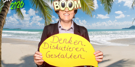 Sommerfest 2026 Dekanin Foto der Dekanin Frau Dr. Ramona Schröpf vor einem tropischen Fotohintergrund mit einem Schild in der Hand mit der Aufschrift "Denken. Diskutieren. Handeln." zum Sommerfest 2026.