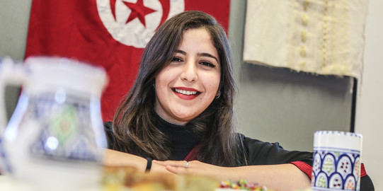 Eine internationale Studierende lächelt in die Kamera. Im Hintergrund hängt eine Tunesienflagge.