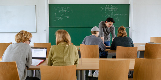 Studierende Tutorium Panorama Vier Studierende im Seminarraum sitzen und arbeiten zusammen mit dem Dozierenden im Hintergrund.