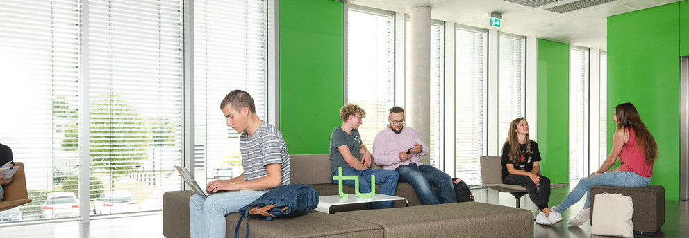 Studierende in einem Gebäude der Uni Sechs Studierende sitzen in einem Aufenthaltsbereich in einem Uni-Gebäude. Sie lesen Bücher, arbeiten am Laptop oder unterhalten sich.