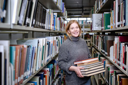 Studierende steht mit einem Stapel Bücher in den Händen zwischen den Bücherregalen der Bibliothek.