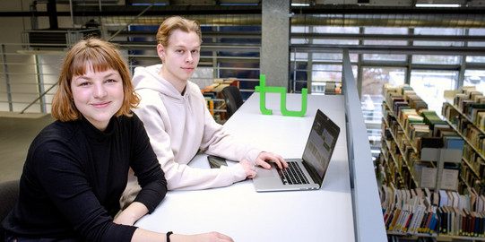 Zwei Studierende sitzen an einem langen Tisch vor einem Laptop in der Bibliothek.