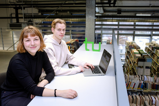 Zwei Studierende sitzen an einem langen Tisch vor einem Laptop in der Bibliothek.