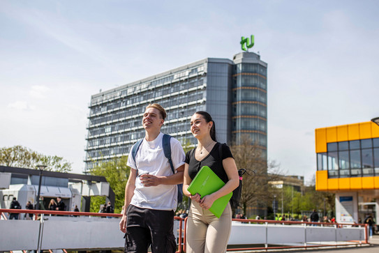 Studierende auf der Mensabrücke Ein junger Mann und eine junge Frau laufen über die Mensabrücke. Im Hintergrund sieht man den Mathetower mit TU-Logo und das Mensagebäude.