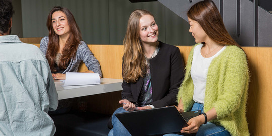 Zwei Studierende unterhalten sich und sitzen gemeinsam auf einer Bank mit einem Laptop.