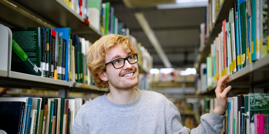Studierender steht suchend an einem Bücherregal in der Bibliothek.