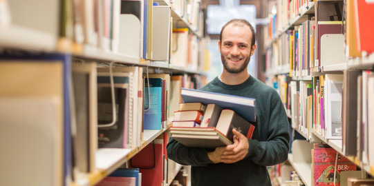 Ein Student steht in der Bibliothek im Gang und hat Bücher auf dem Arm.