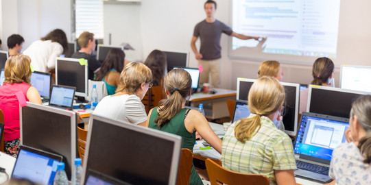 Viele Menschen sitzen in einem Kursraum vor Computern. Vorne steht ein Lehrer und zeigt etwas auf einem Bildschirm.