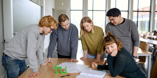Studierende Gruppenarbeit Fünf Studierende im Seminarraum stehen um einen Tisch herum und sehen auf ein Dokument