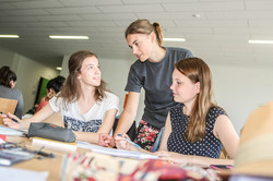 Zwei Personen sitzen mit Unterlagen und Stiften an einem Tisch. Eine dritte Person beugt sich zwischen die beiden Personen und spricht mit der Person, die links sitzt. Im Hintergrund sieht man weitere Personen an Tischen.