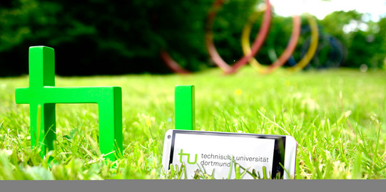 Ein Smartphone, das neben dem TU-Logo im Gras steht, im Hintergrund sind die Spektralringe zu sehen.