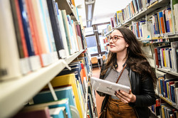 Studierende steht mit einem Tablet in der linken Hand an den Bücherregalen in der Bibliothek und sucht ein Buch.