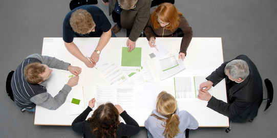 Studierende und Dozierende sitzen gemeinsam an einem Tisch und arbeiten.