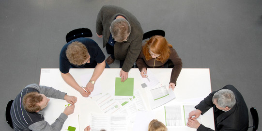 Studierende und Dozierende sitzen gemeinsam an einem Tisch und arbeiten.