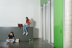 Eine Studentin sitzt auf dem Boden und liest, eine andere auf einer Theke und liest. Rechts im Bild ein großes Fenster.
