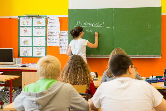 in einem Klassenzimmer sitzen Schüler*innen, eine junge Frau schreibt an der Tafel
