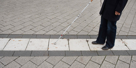 Eine blinde Person folgt dem Blindenleitsystem auf dem Campus mithilfe eines Blindenstocks