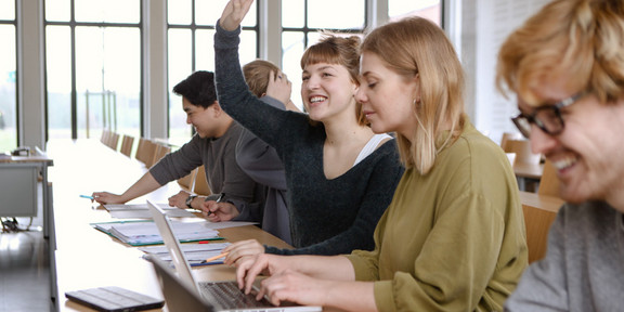 Fünf Studierende sitzen in einem Seminarraum an Tischen. Eine Studentin hebt ihre Hand.