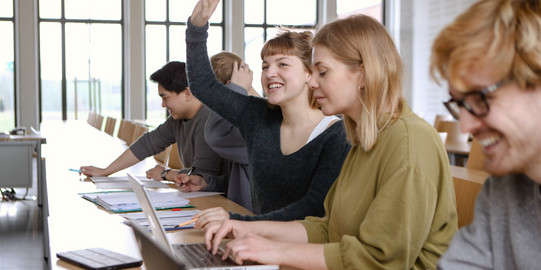 Studierende im Seminar Fünf Studierende sitzen in einem Seminarraum an Tischen. Eine Studentin hebt ihre Hand.