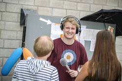 Ein Student mit Kopfhörern auf dem Kopf und Mikrofon in der Hand unterhält sich mit einem Studenten und einer Studentin, die an seinem Stand stehen.